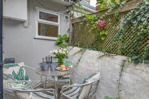An outdoor patio with a table set for two, surrounded by metal chairs with cushions. A small table holds a pitcher, cups, and a plate with food. - 2 Bed in Padstow (oc-p00821) (Padstow)