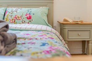 Ein Bett mit floraler Bettwäsche, ein Nachttisch mit einem Buch und einer Tasse, sowie ein Beistelltisch mit einer Schublade.