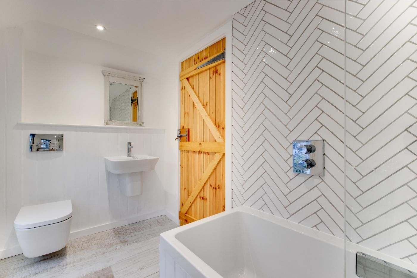 Une salle de bain avec un lavabo, des toilettes et une baignoire blancs, une porte en bois et un mur orné d’un motif en chevrons.