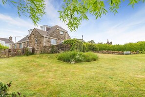 Une maison en pierre avec un toit en pente, une cour clôturée et un petit arbuste au premier plan.