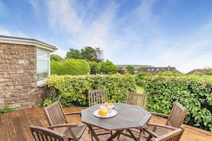 Une terrasse en bois avec une table et des chaises, entourée de haies et d’un mur de pierres.