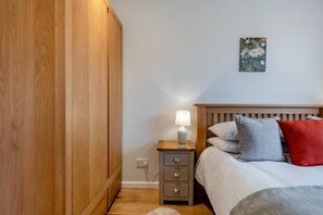 A bedroom with a wooden wardrobe, a bedside table with a lamp, a bed with pillows, and a wall-mounted painting. - 3 Bed in Worth Matravers (oc-h32107) (Worth Matravers)