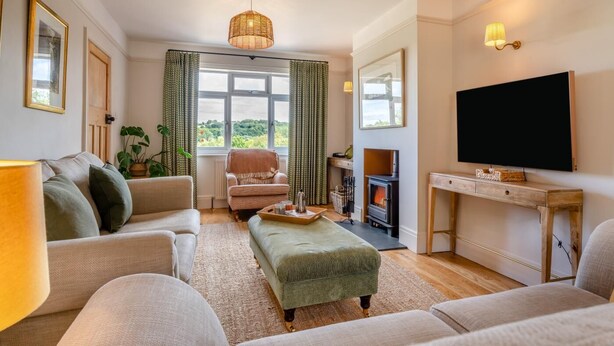 A cozy living room with a sofa, armchair, and ottoman, a fireplace, a television, and a large window with curtains. - 4 Bed in Ruswarp (oc-d32269) (Ruswarp)