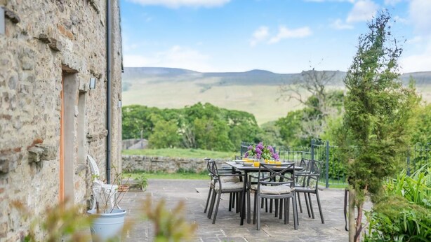 A stone building with a table and chairs set outdoors, surrounded by greenery and a view of hills. - 4 Bed in Burtersett (oc-h32270) (Burtersett)