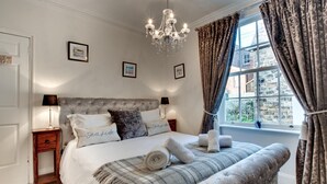 A bedroom with a tufted headboard, a large bed with white linens, two bedside tables with lamps, a chandelier, and a view of a brick building through a window with curtains. - 4 Bed in Whitby (oc-w474) (Whitby)