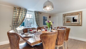 A dining room with a wooden table set for six, surrounded by six leather chairs with bead trim. A large mirror hangs on the wall, and a window with plaid curtains is visible. - 4 Bed in Whitby (oc-w474) (Whitby)