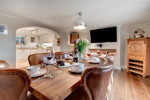 A dining area with a wooden table set for six, surrounded by leather chairs. A television is mounted on the wall, and a wooden cabinet with wine bottles is visible. - 4 Bed in Whitby (oc-w474) (Whitby)