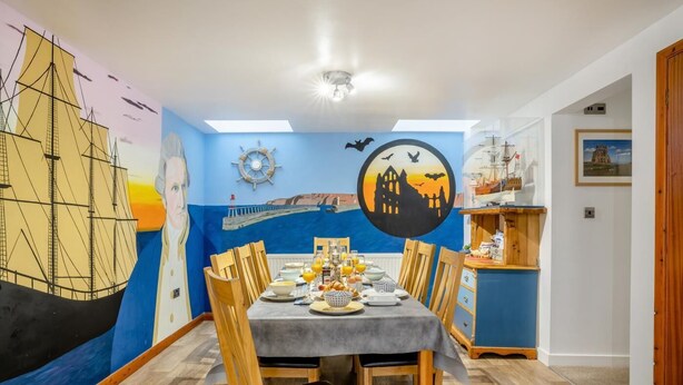 Uma área de refeições com um mural de um navio e pôr do sol, cadeiras de madeira, uma mesa posta para o café da manhã e uma pequena prateleira de madeira com objetos.