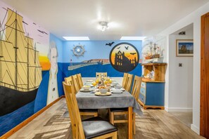 A dining area with a mural of a ship and sunset, wooden chairs, a table set with breakfast, and a small wooden shelf with items. - 4 Bed in Whitby (oc-w506) (Whitby)