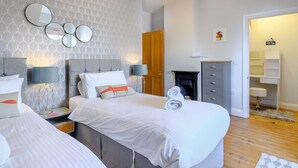 Een moderne slaapkamer met behang met een grijs patroon, een gestoffeerd grijs hoofdbord, twee nachtkastjes met lampen, een grijze dressoir en een houten deur.
