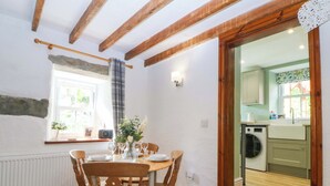 A dining area with a table set for four, wooden chairs, a window with curtains, and a view into a kitchen with a washing machine. - Old Chapel House (Llanfairfechan)