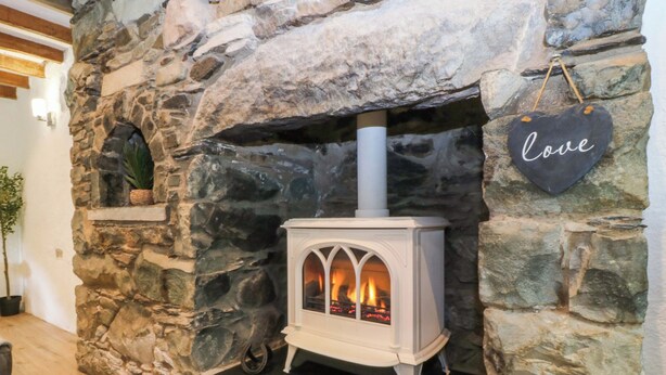 A stone fireplace with a white wood-burning stove, a hanging 'love' sign, and a potted plant on the left. - Old Chapel House (Llanfairfechan)