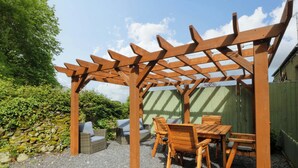 A wooden pergola with a table and chairs underneath, surrounded by a gravel area and greenery. - Old Chapel House (Llanfairfechan)