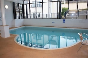 Piscina interior com paredes de vidro transparente, chão ladrilhado e guarda-corrimão de metal.
