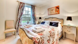 Ein Schlafzimmer mit einem großen Bett, floralen Vorhängen, einer Chandelier, einem Gemälde und einem Nachttisch mit Lampe.