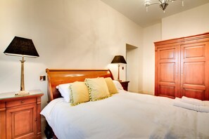 Ein ordentlich bezogenes Bett mit Kissen, ein Nachttisch aus Holz mit einer Lampe, ein Kleiderschrank und eine Wandlampe.