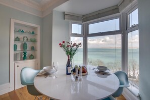 A dining area with a round table, chairs, a vase with flowers, and a view of the sea through large windows. - 1 Bed in St. Ives (oc-aqua19) (St Ives)