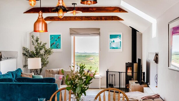 A cozy living room with a blue sofa, a dining table with chairs, a fireplace, and a view of the countryside. - 3 Bed in Mawgan Porth (oc-p00824) (Mawgan Porth)
