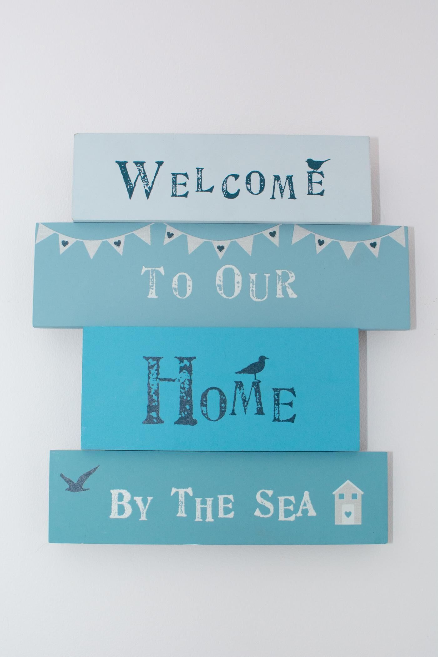 Un cartel de bienvenida con el texto 'WELCOME TO OUR HOME By The Sea' y elementos decorativos como pájaros y un icono de una casa.