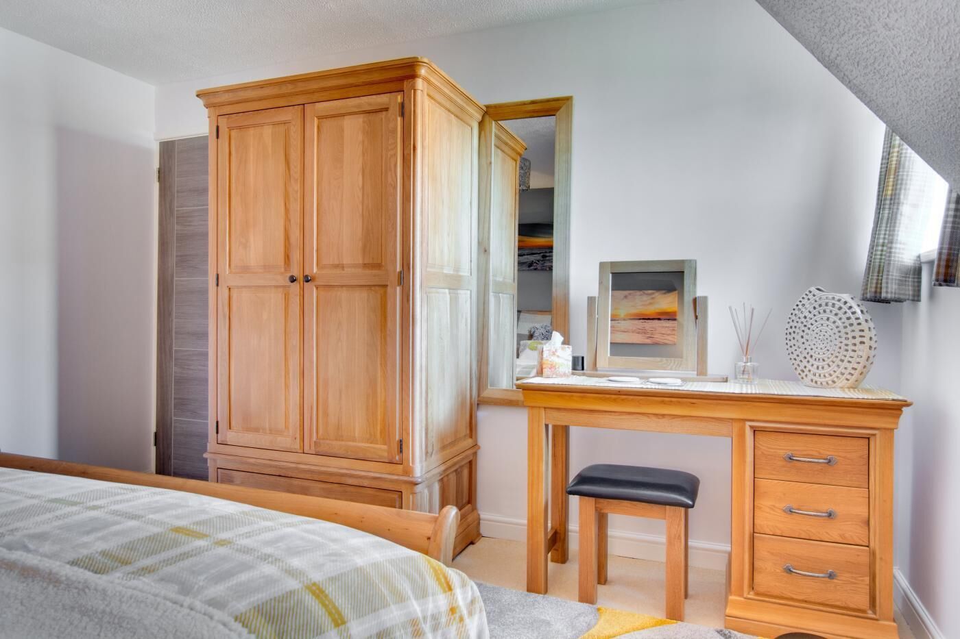Een slaapkamer met een houten kledingkast, een bureau met een spiegel, een klein krukje en een bed met een gestreept dekbed.
