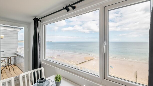 Habitación con un ventanal grande que da a una playa, una mesa de comedor con platos y una planta en maceta, y vista al océano.