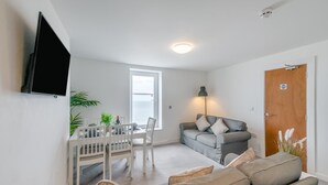 A living room with a grey sofa, a dining table with chairs, a TV mounted on the wall, and a door with a sign. - 3 Bed in Bridlington (oc-o32293) (Bridlington)