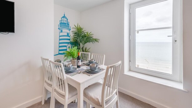 A dining area with a white table set for four, white chairs, a potted plant, and a bottle of wine. A large window offers a view of the sea. - 3 Bed in Bridlington (oc-o32293) (Bridlington)
