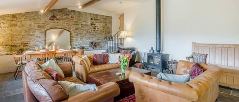 A cozy living room with leather sofas, a stone wall, a wooden staircase, and a wood-burning stove.
