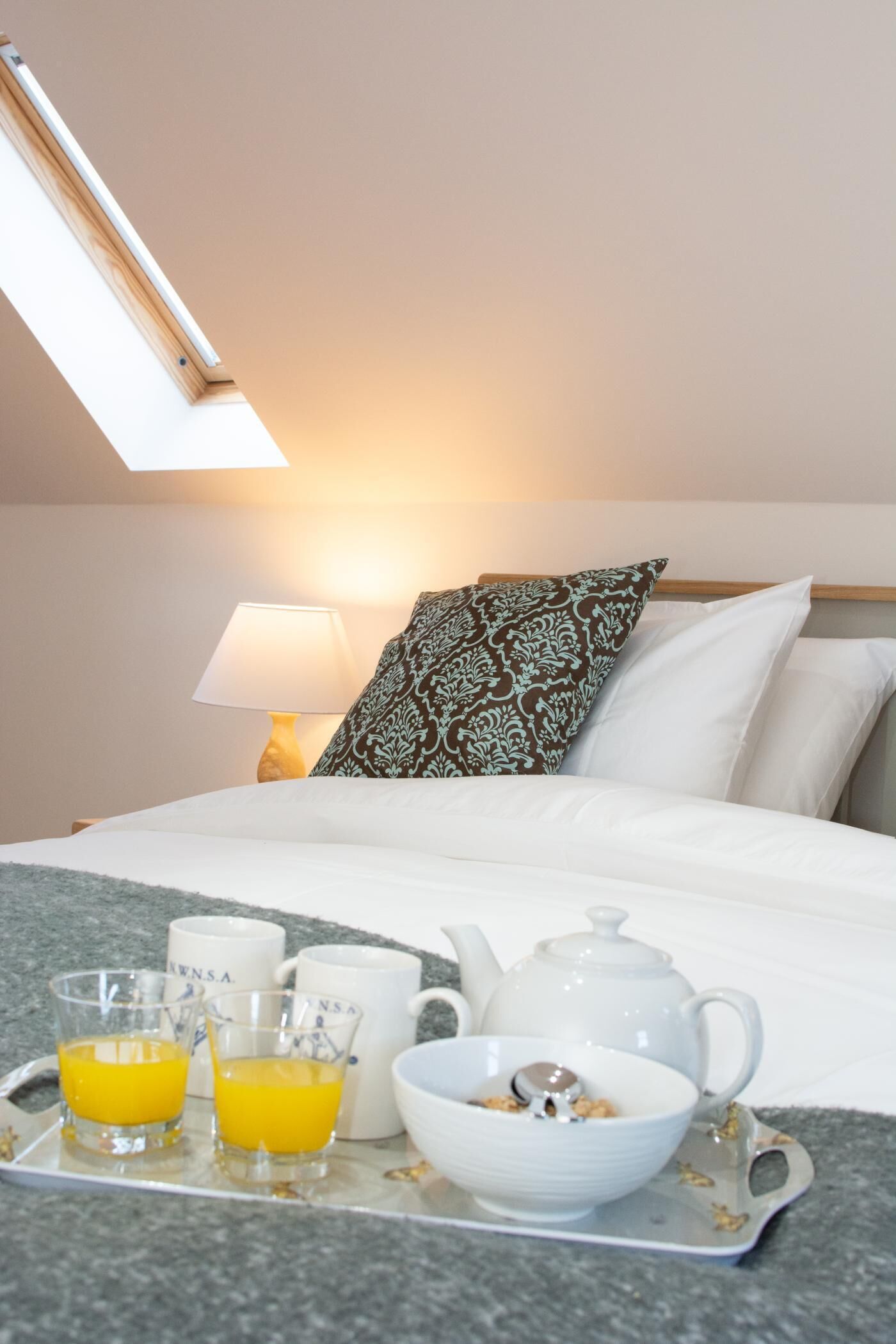 A neatly made bed with white linens, a patterned pillow, a bedside lamp, and a tray with breakfast items including orange juice and a bowl of cereal.