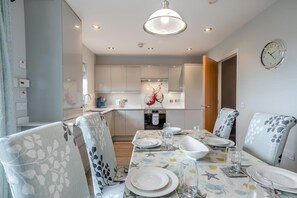 A modern dining area with a table set for four, a kitchen in the background, and a clock on the wall. - 4 Bed in Amble (oc-gd1868) (Amble)