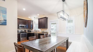 A dining area with a wooden table and chairs, a kitchen with modern appliances, and a view of the outside through a door. - 3min Walk to Beach with Heated Pool in Ocean Sands (Corolla)