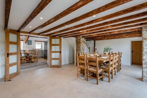 A spacious dining area with a high wooden ceiling and exposed beams, a stone wall, and a view into a kitchen area. - 5 Bed in Nenthead (oc-f32234) (Alston Moor)