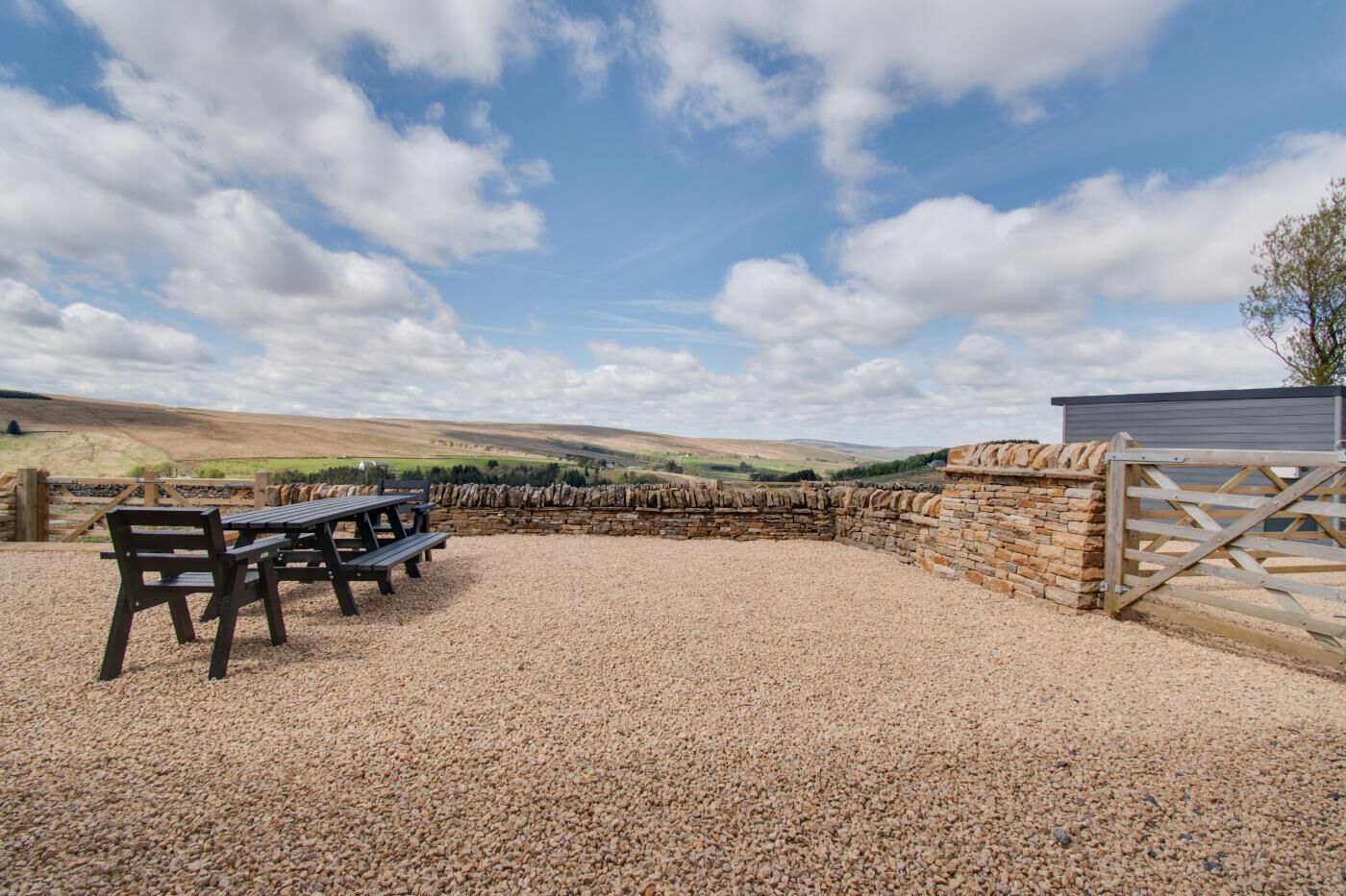 A gravel patio with picnic tables, a stone wall, and a wooden gate.