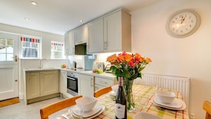 A modern kitchen with light grey cabinets, a dining table set with white plates, bowls, and a wine bottle. A vase of flowers is on the table, and a wall clock is visible. - 3 Bed in St Ives (oc-vgn25) (St Ives)