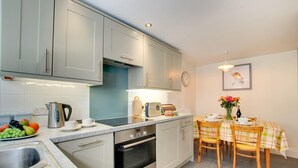 A modern kitchen with stainless steel appliances, a dining area with a table set for two, and a clock on the wall. - 3 Bed in St Ives (oc-vgn25) (St Ives)