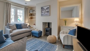 A living room with a sofa, ottoman, and armchair, a fireplace, a television, and a large mirror. - 3 Bed in St Ives (oc-vgn25) (St Ives)