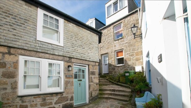 A narrow stone pathway leading to a cottage with a light blue door, a small staircase, and a white building with a window and a lamp post. - 3 Bed in St Ives (oc-vgn25) (St Ives)