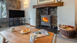 A cozy living room with a stone wall, a fireplace, a television, and a table with pastries and a magazine. - 1 Bed in Llanfair (oc-b32109) (Llanfair)