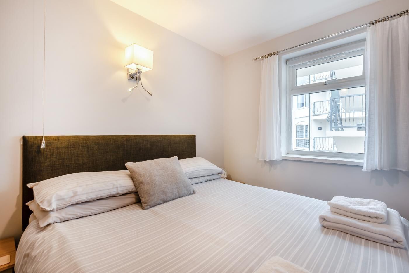 A neatly made bed with white linens and a single pillow, a wall-mounted light fixture, and a window with curtains.