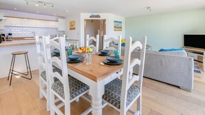 A modern kitchen with white cabinets, a wooden dining table with white chairs, and a grey sofa in the living area. - 3 Bed in Woolacombe (oc-c32222) (Woolacombe)