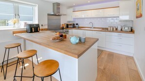 A modern kitchen with a central island, bar stools, a stove, microwave, and white cabinetry. - 3 Bed in Woolacombe (oc-c32222) (Woolacombe)