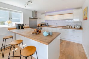 A modern kitchen with a central island, bar stools, a stove, microwave, and white cabinetry. - 3 Bed in Woolacombe (oc-c32222) (Woolacombe)