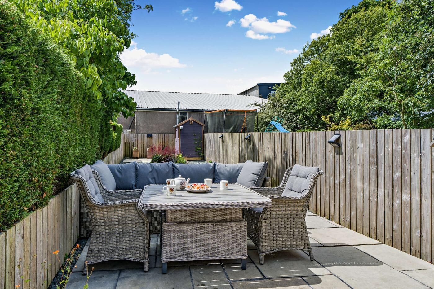 A patio with wicker furniture and a table set for two, surrounded by a wooden fence and greenery.