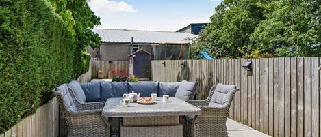 A patio with wicker furniture and a table set for two, surrounded by a wooden fence and greenery.