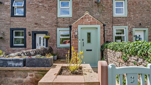 Una casa de piedra con varias ventanas, una puerta verde y un pequeño jardín con plantas y un banco.