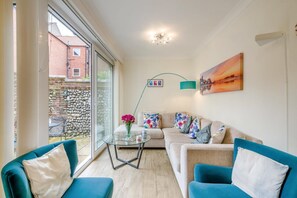 A modern living room with a sofa, two armchairs, a glass coffee table, and a view of a stone wall and building outside. - 3 Bed in Cromer (oc-2103) (Cromer)