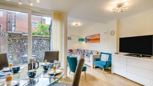 A modern living room with a glass dining table, a sofa, a TV, and a view of a brick wall and greenery outside. - 3 Bed in Cromer (oc-2103) (Cromer)