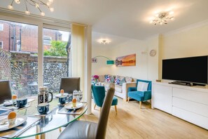 A modern living room with a glass dining table, a sofa, a TV, and a view of a brick wall and greenery outside. - 3 Bed in Cromer (oc-2103) (Cromer)