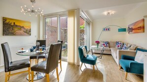 A modern living room with a glass dining table, comfortable seating, a sofa, and a view through large windows. - 3 Bed in Cromer (oc-2103) (Cromer)