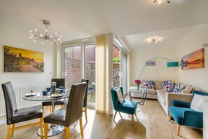 A modern living room with a glass dining table, comfortable seating, a sofa, and a view through large windows. - 3 Bed in Cromer (oc-2103) (Cromer)